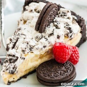 A slice of Oreo banana cream cake on a white plate with a fork, garnished with an oreo cookie and two raspberries.
