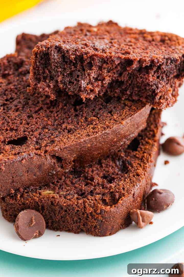 Decadent Chocolate Banana Bread 5 Two and a half slices of beautifully baked chocolate banana bread, stacked on a white plate, with the top slice gently broken to reveal its moist interior, garnished with chocolate chips.