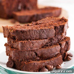 A stack of four moist slices of chocolate banana bread, garnished with chocolate chips, on a white plate, with the full loaf blurred in the background.