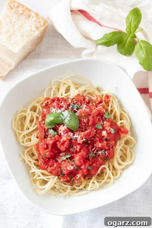 Homemade Fresh Tomato Pasta Sauce with Basil and Parmesan A white plate with spaghetti topped generously with homemade fresh tomato sauce, garnished with bright green basil leaves and a dusting of grated Parmigiano cheese. Next to the plate are fresh basil sprigs, a white dish towel with a red stripe, and a block of Parmigiano cheese, highlighting the key ingredients.