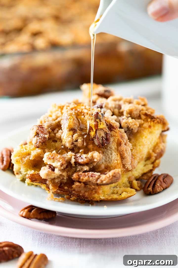 A slice of golden-brown Overnight French Toast Casserole with a generous topping of crunchy pecans and streusel, served on a white plate. Maple syrup cascades elegantly over the warm breakfast. In the background, a full glass baking dish of the casserole rests, hinting at more deliciousness to come.