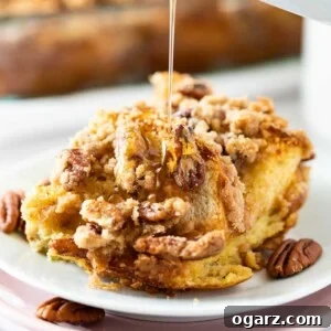 A piece of french toast casserole with pecans and streusel on a white plate stacked on a pink plate garnished with pecans. A small white jar is pouring maple syrup over it. There's a glass baking dish with the casserole in the background.