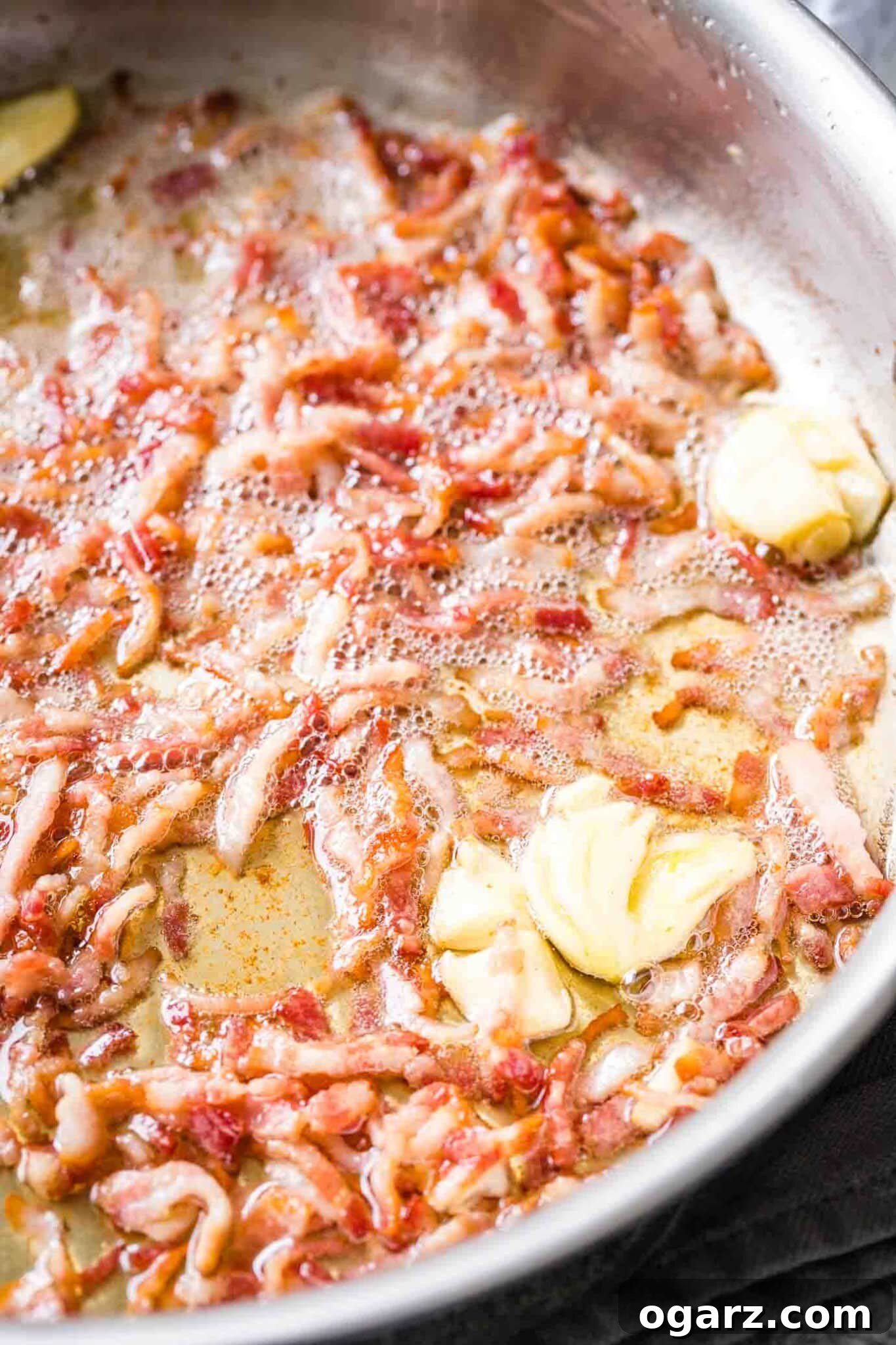 Pasta Carbonara is an easy Italian recipe that can be made in less than 20 minutes from start to finish and is so creamy and delicious. This classic carbonara sauce can be served with your favorite kind of pasta like spaghetti or fettuccine and makes an indulgent and quick weeknight dinner! #pastacarbonara #pastarecipes #spaghetti Bacon, cut into small pieces, being fried in a stainless tell pan, with some crushed and peeled garlic cloves.