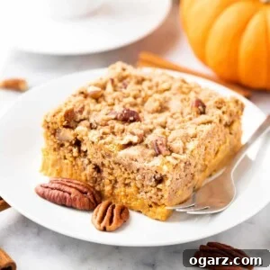 A piece of pumpkin cake with pecans on a white plate with a fork, garnished with whole pecans, cinnamon sticks and pumpkin. There's a cup of espresso in the background