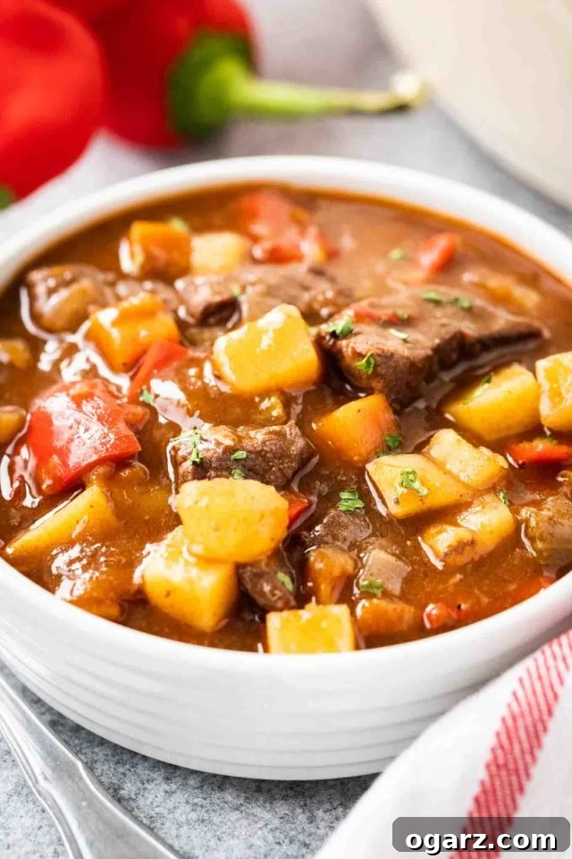 Hearty Hungarian Goulash: Tender Beef and Paprika Perfection A white bowl of goulash, containing red peppers, beef and potatoes, topped with fresh parsley. There's a white dish towel with red stripes and some red peppers next to it.
