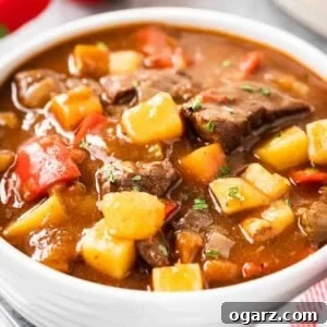 A white bowl of goulash, containing red peppers, beef and potatoes, topped with parsley. There's a white dish towel with red stripes and some red peppers next to it.