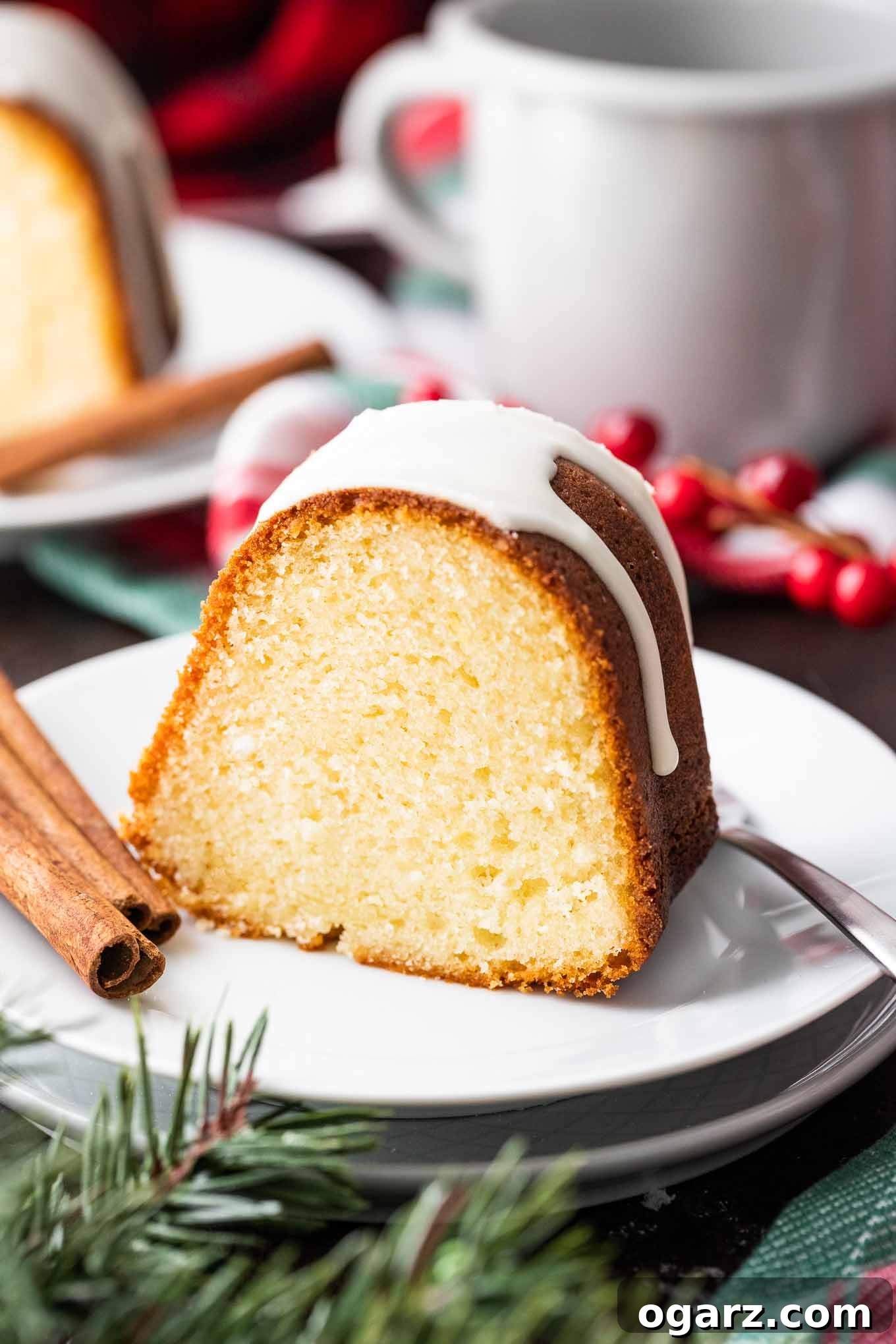 Moist Eggnog Bundt Cake with Glaze: The Perfect Make-Ahead Holiday Dessert #EggnogCake #HolidayCake #BundtCake #HolidayDesserts A slice of perfectly glazed Eggnog Bundt Cake on a white plate, garnished with fresh cranberries and cinnamon sticks. A steaming cup and more festive decor are blurred in the background, creating a cozy holiday ambiance.