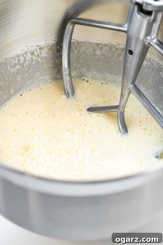 Fluffy Eggnog Cake Batter Ready for Baking #BakingTips #CakeBatter #StandMixer A close-up shot of light yellow cake batter in a stainless steel stand mixer bowl, with the flat beater attachment partially submerged, illustrating the ideal texture for baking.