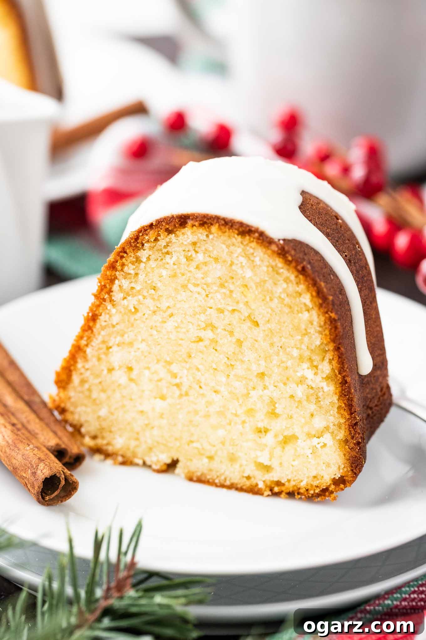 Delicious Slice of Eggnog Cake with Glaze and Festive Garnishes #HolidayDessert #EggnogFlavor #FestiveBaking A close-up view of a single slice of Eggnog Bundt Cake, generously drizzled with a rich white glaze, presented elegantly on a white plate alongside a fork, two cinnamon sticks, and scattered cranberries. The background features a cozy, festive setting with a cup and another plate, emphasizing the cake's holiday appeal.