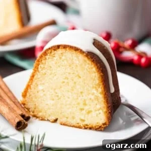 A piece of bundt cake with white glazing on top on a white plate with a fork and two sticks of cinnamon. There are cranberries, a cup and another plate in the background