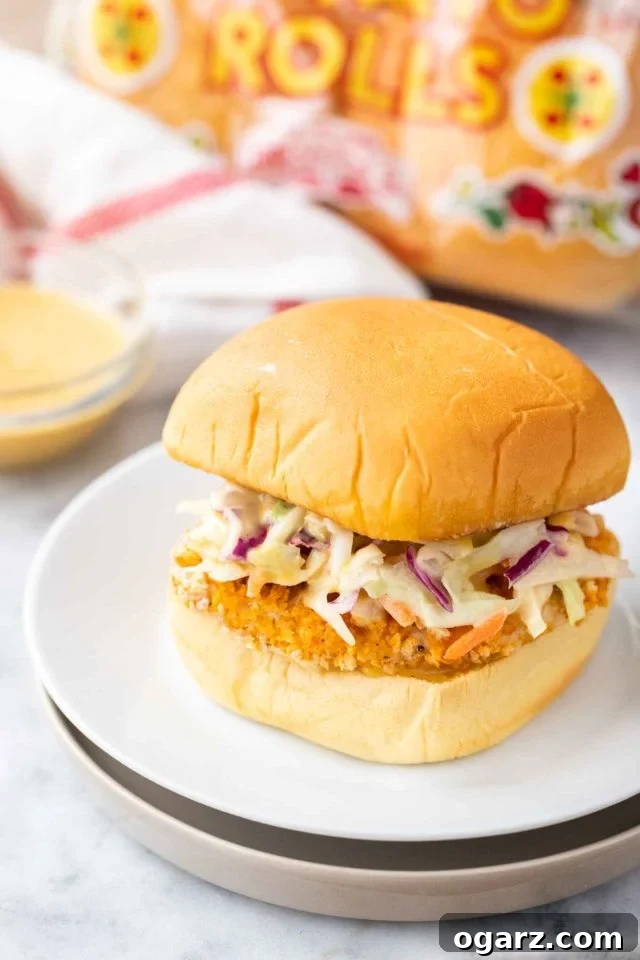Crispy Air Fried Chicken Sandwiches 2 A burger with a breaded fried chicken patty and coleslaw on a white plate next to a glass bowl with dip, a white and red towel and a bag of burger buns