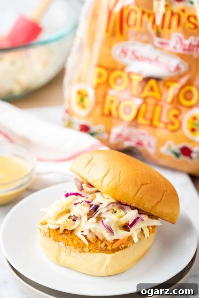 Crispy Air Fried Chicken Sandwiches 4 A burger with a breaded fried chicken patty and coleslaw with the top sliding off on a white plate next to a glass bowl with dip, a white and red towel, a glass bowl with coleslaw and a bag of burger buns