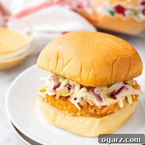 A burger with a breaded fried chicken patty and coleslaw on a white plate next to a glass bowl with dip, a white and red towel and a bag of burger buns