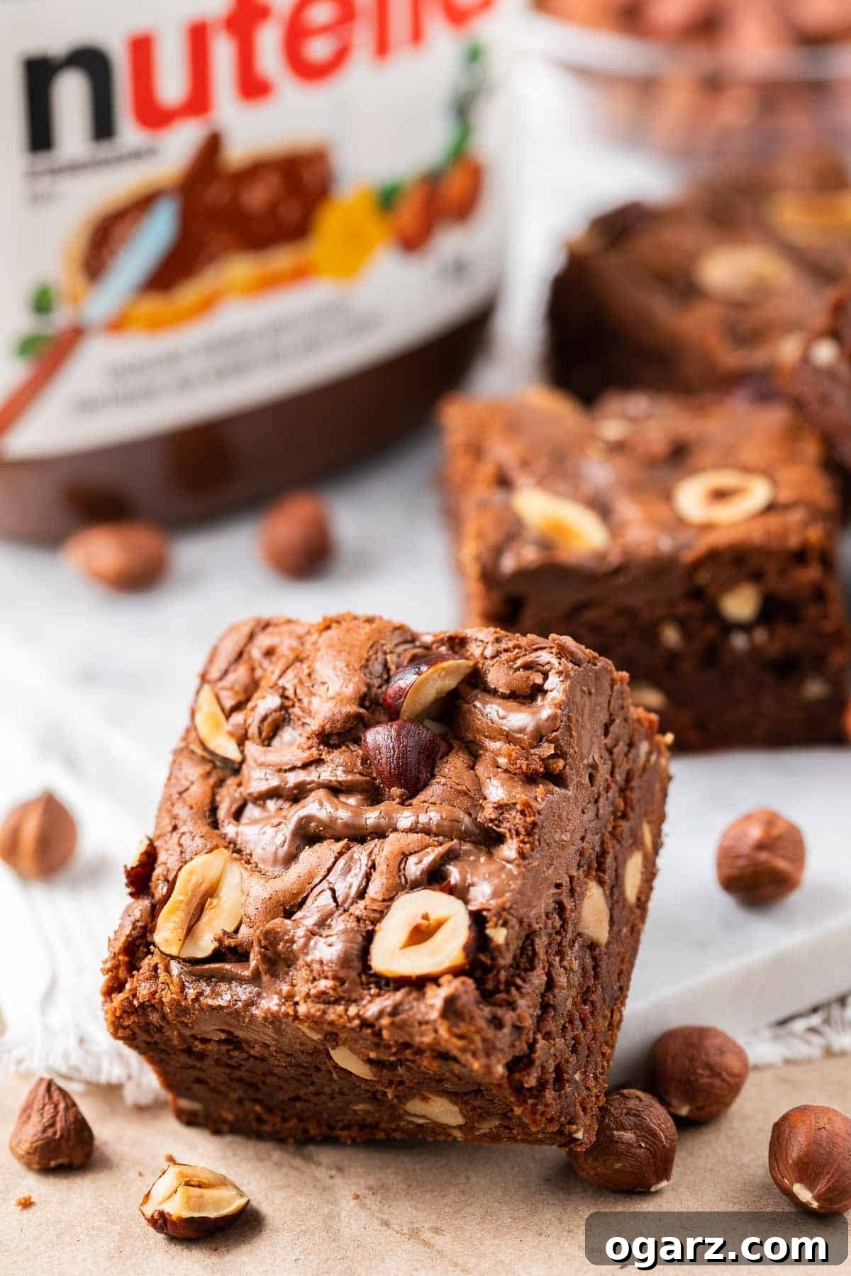 Fudgy Nutella brownie leaning against a white plate, surrounded by whole hazelnuts, a jar of Nutella, and more delicious Nutella brownies in the background.