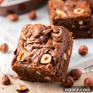 Fudgy Nutella brownie leaning against a white plate with hazelnuts, a jar of Nutella and several Nutella brownies in the background