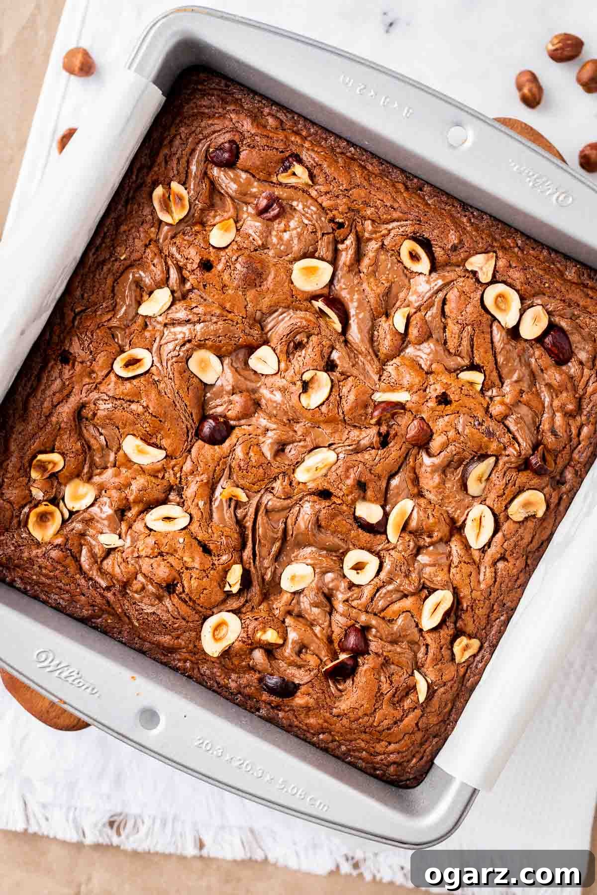 A perfectly baked pan of Nutella brownies, still warm, with visible Nutella swirls and a generous sprinkling of crunchy hazelnuts, ready to be cooled and sliced.