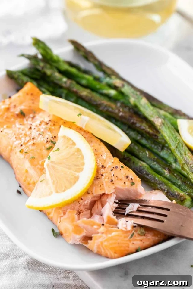 A beautifully plated dish featuring a flaky, cooked salmon fillet next to vibrant roasted green asparagus, garnished with fresh lemon slices. A bronze fork is gently flaking the salmon to show its perfect tenderness.