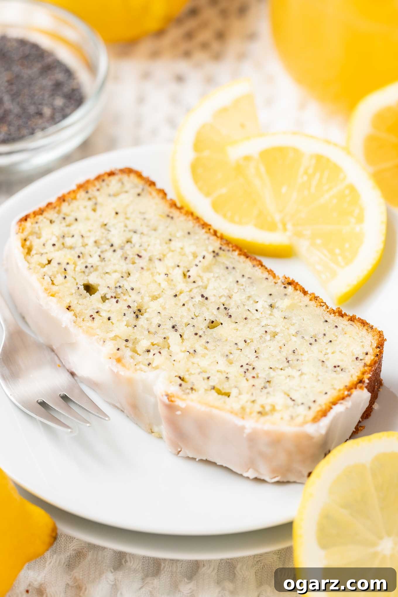 Bright Lemon Poppy Seed Bread 4 A slice of lemon poppy seed cake on a white plate with a fork, garnished with lemon slices and a glass bowl with poppy seeds in the background.