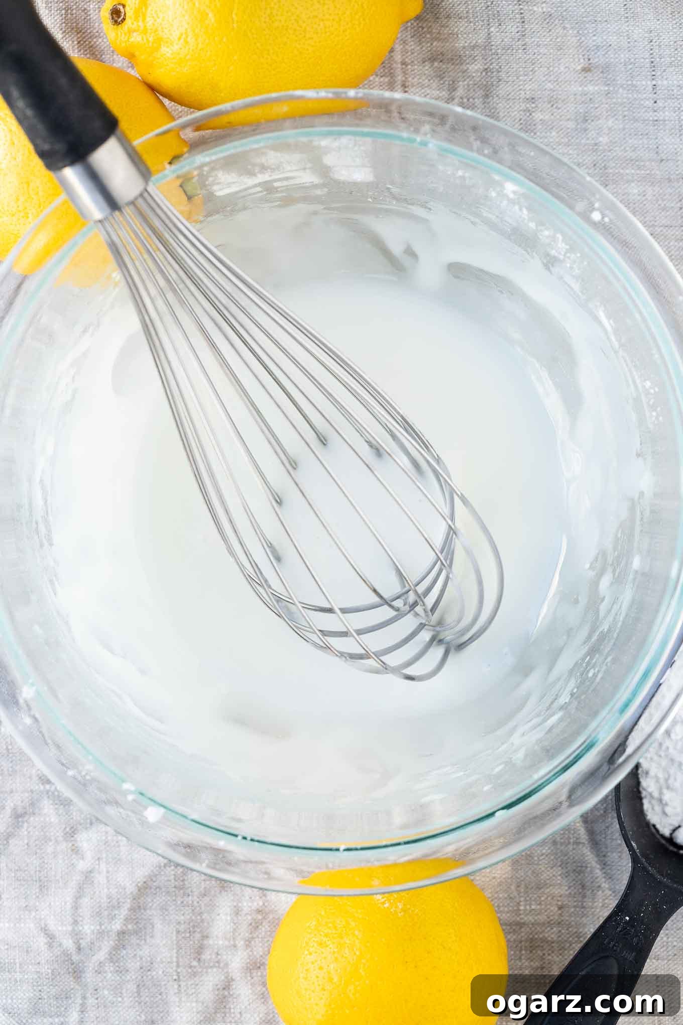 Bright Lemon Poppy Seed Bread 5 Lemon glaze in a glass bowl with a whisk in it and a lemon and a bowl of powdered sugar next to it.