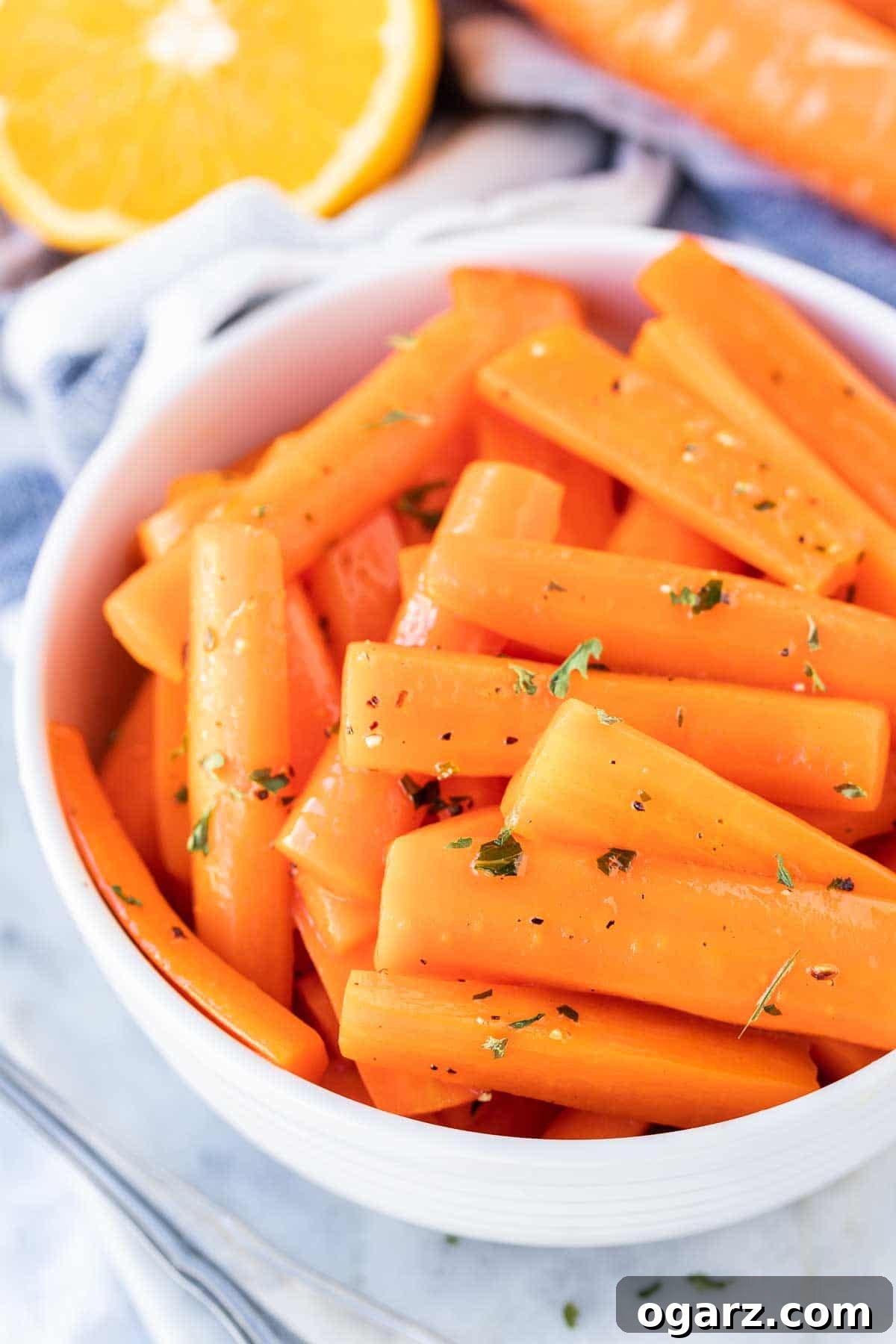 A bowl of glossy honey glazed carrots, garnished with fresh parsley and ready to be enjoyed as a festive or everyday side dish. Glazed Carrots in a serving bowl with an orange next to it, garnished with parsley.