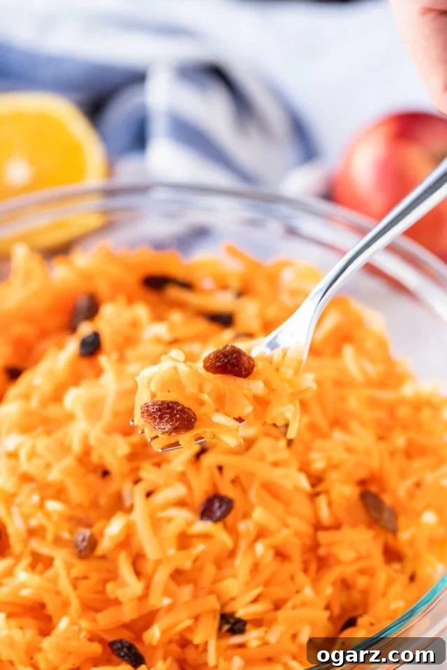 This easy Carrot Salad with Raisins and Apple is a bright and colorful side dish that adults and kids will love! Made with shredded carrots and apples this delicious German Carrot Raisin Salad Recipe can be made in minutes and is loaded with healthy vitamins. #CarrotRaisinSalad #CarrotSalad #SaladRecipes Carrot salad with raisins in a glass bowl, with a portion of it on a fork hovering above it. Half and orange and an apple in the background