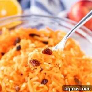 Zesty Carrot Salad 2 Carrot salad with raisins in a glass bowl, with a portion of it on a fork hovering above it. Half and orange and an apple in the background