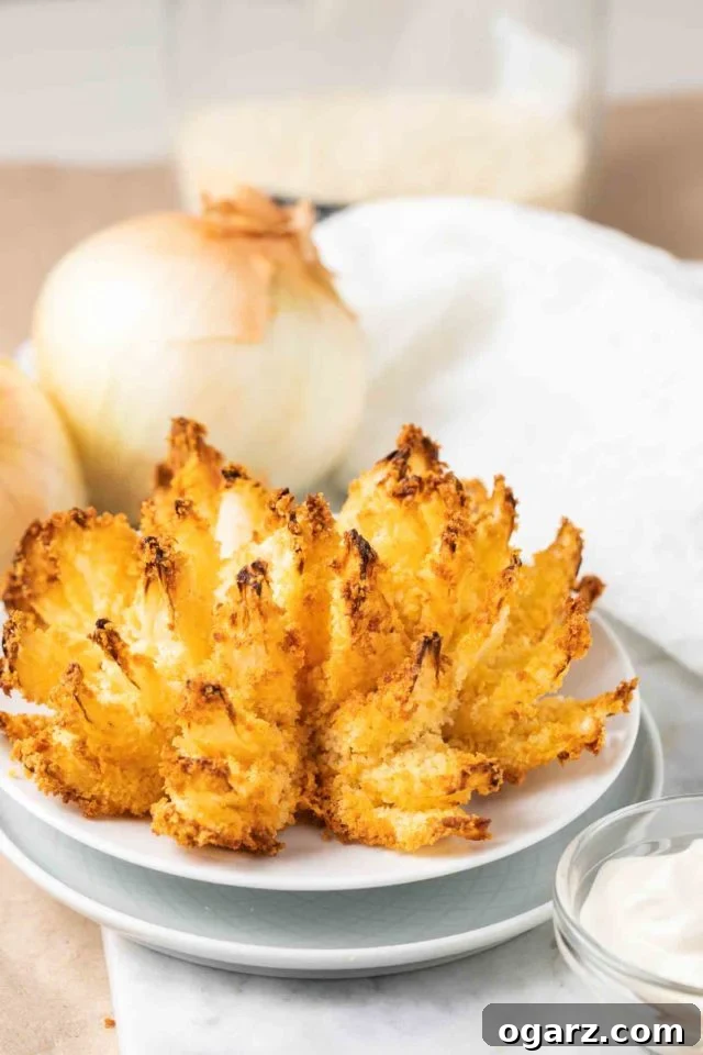 Air Fried Onion Blossom 2 Golden brown and perfectly crispy Air Fryer Blooming Onion on a white serving plate, with blurred whole onions in the background, highlighting its appeal as a gourmet appetizer.