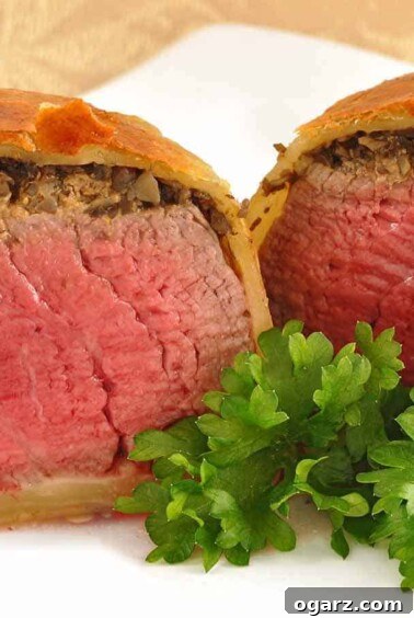 A dinner plate showcasing an individual Beef Wellington, expertly sliced in half to reveal perfectly cooked beef tenderloin, a savory mushroom duxelles topping, and a beautifully flaky puff pastry wrap.