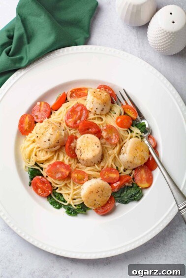 Delicate Scallops and pasta with a luminous white wine butter sauce, served over fine angel hair pasta with vibrant grape tomatoes and tender sautéed spinach on a white plate.