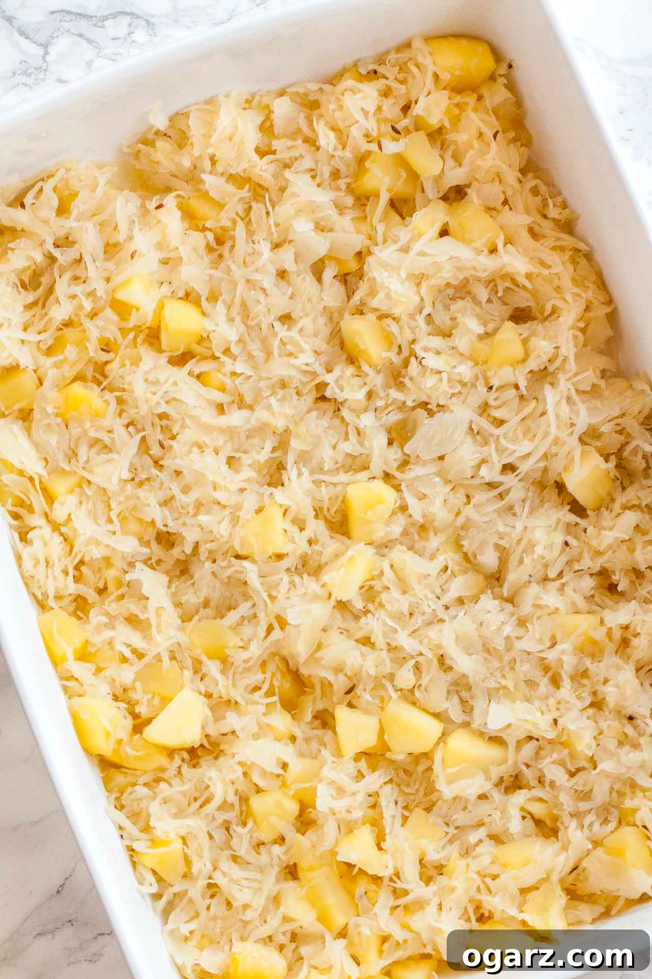 Tangy Kraut and Creamy Potato Bake 4 A baking dish containing a vibrant layer of chopped apple pieces and seasoned sauerkraut, ready for the mashed potato topping, displayed on a sleek marble surface.