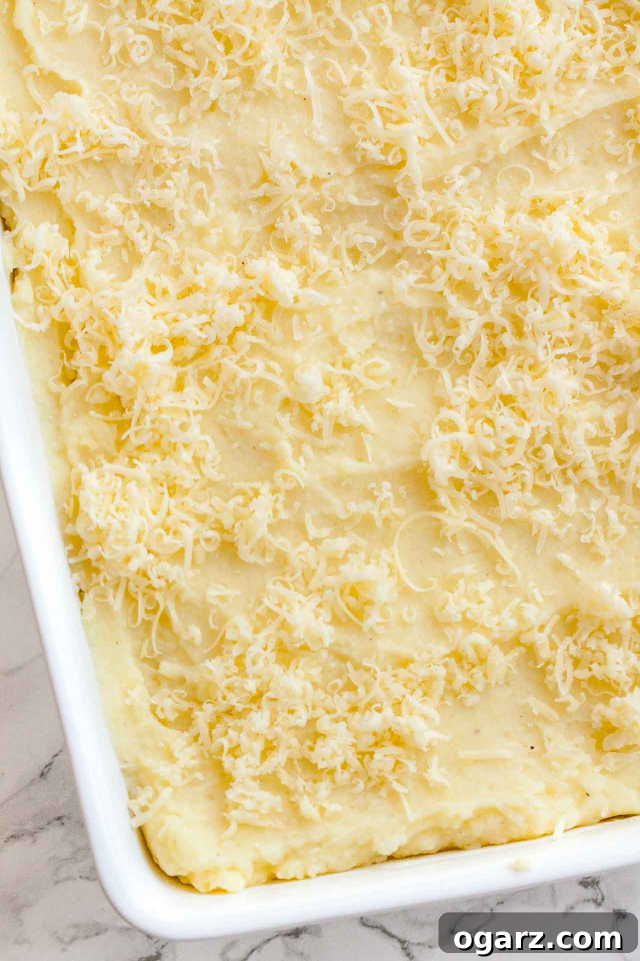 Tangy Kraut and Creamy Potato Bake 5 A close-up of a white baking dish filled with a creamy layer of mashed potatoes, generously topped with shredded cheddar cheese, sitting on a pristine marble surface, ready for the breadcrumb topping.