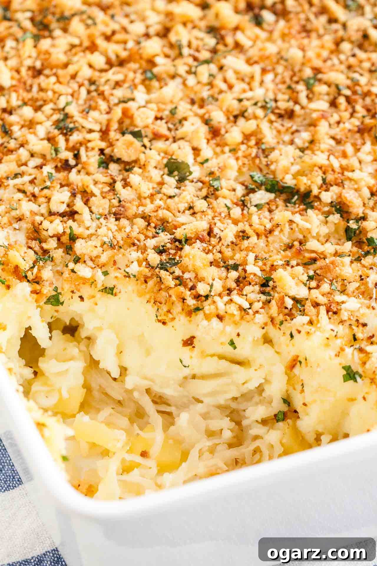 Tangy Kraut and Creamy Potato Bake 7 A portion of the golden-brown Sauerkraut Mashed Potato Casserole has been scooped out, revealing the creamy layers beneath, presented in a white baking dish on a patterned blue and white kitchen towel.