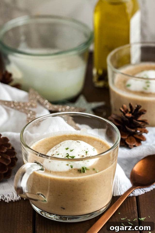 A glass cup of chestnut soup with truffle froth on a wooden table with a bronze spoon next to it. There's a white tablecloth with Christmas ornaments and another cup of soup in the background.
