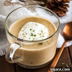 A glass cup of chestnut soup with truffle froth on a wooden table with a bronze spoon next to it. There's a white tablecloth with Christmas ornaments in the background.