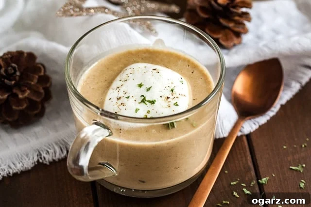 A glass cup of chestnut soup with truffle froth on a wooden table with a bronze spoon next to it. There's a white tablecloth with Christmas ornaments in the background.