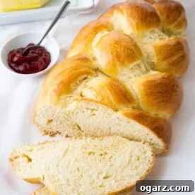 A Hefezopf, with a slice cut off and lying in front of it on a white dish towel next to a small white bowl of strawberry jam with a spoon in it.