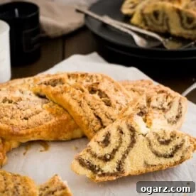 A nut roll, with braided strands of dough with hazelnuts on white parchment paper with a slice cut off and leaning against it. There's a black plate with two forks and more of the nut roll in the background.